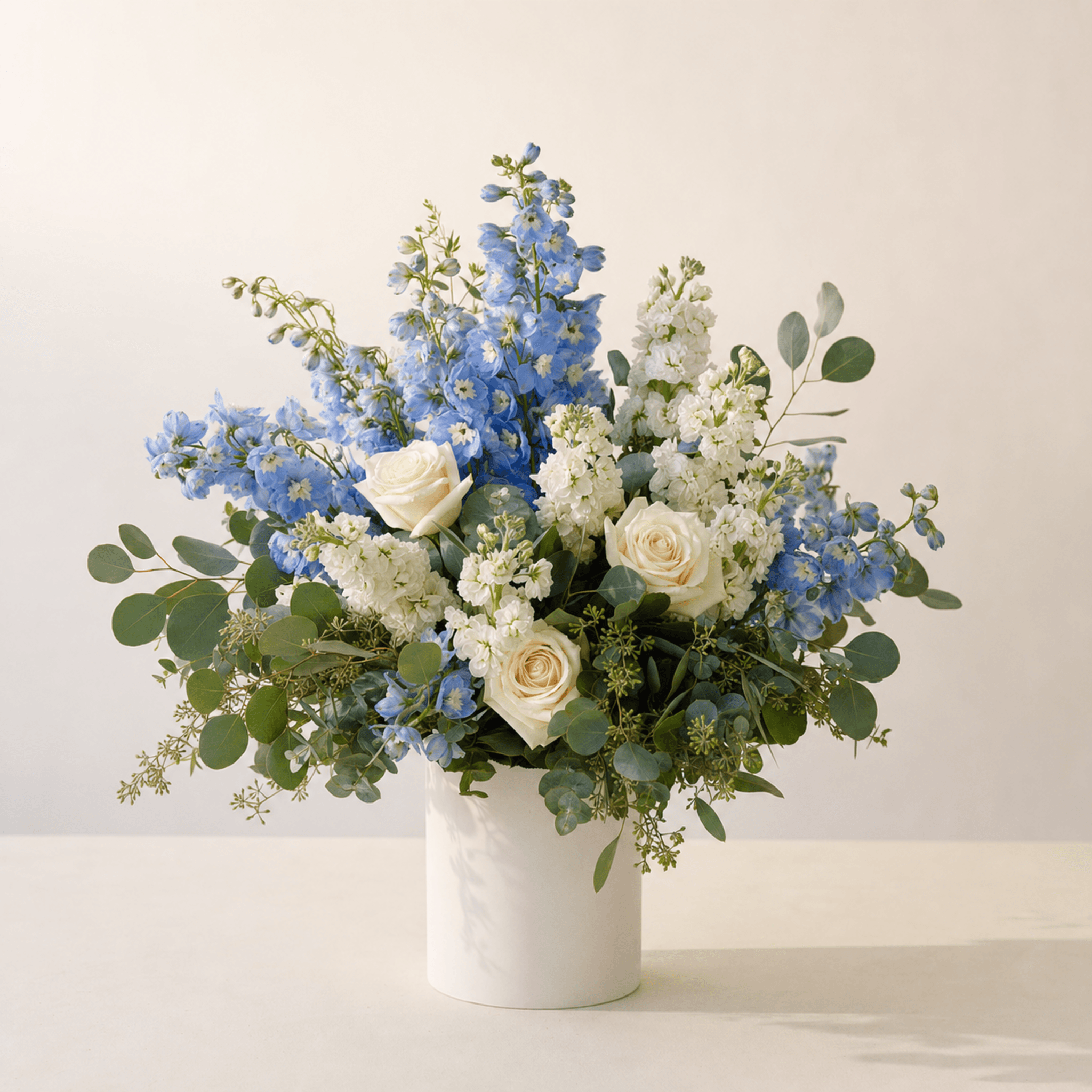 Blue delphinium, white stock, cream roses, and eucalyptus in a white marble cylinder vase