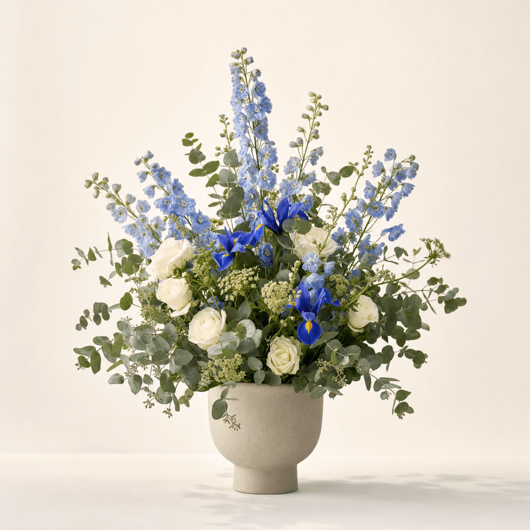 Blue delphinium, iris, white roses, and eucalyptus in a wide ceramic footed vase