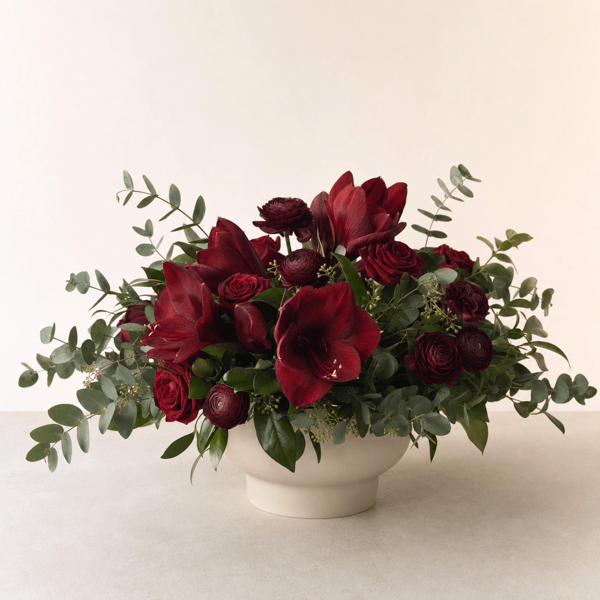 Deep red amaryllis, roses, ranunculus, and eucalyptus in a cream footed ceramic bowl