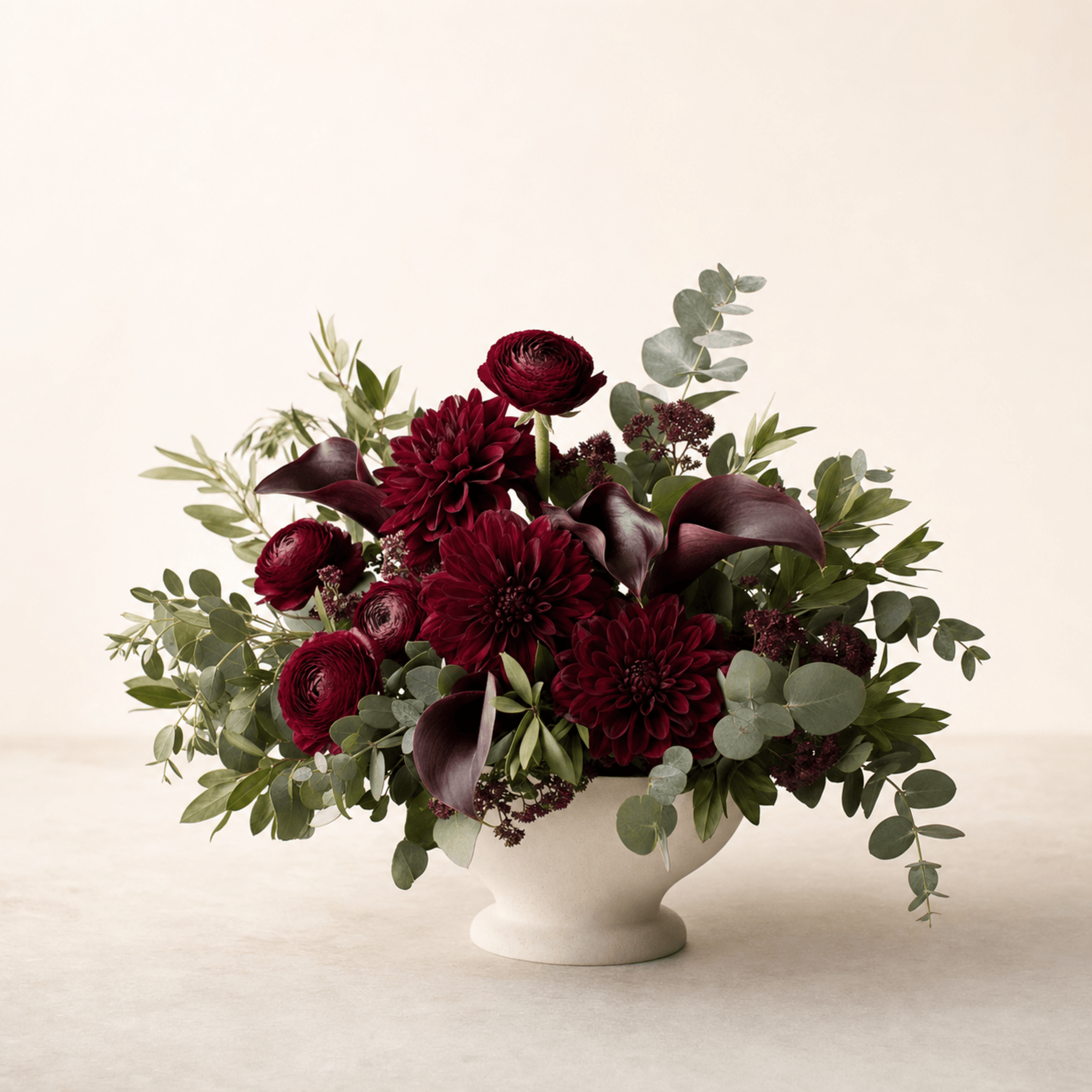 Deep red dahlias, ranunculus, and dark calla lilies with eucalyptus in a white footed bowl