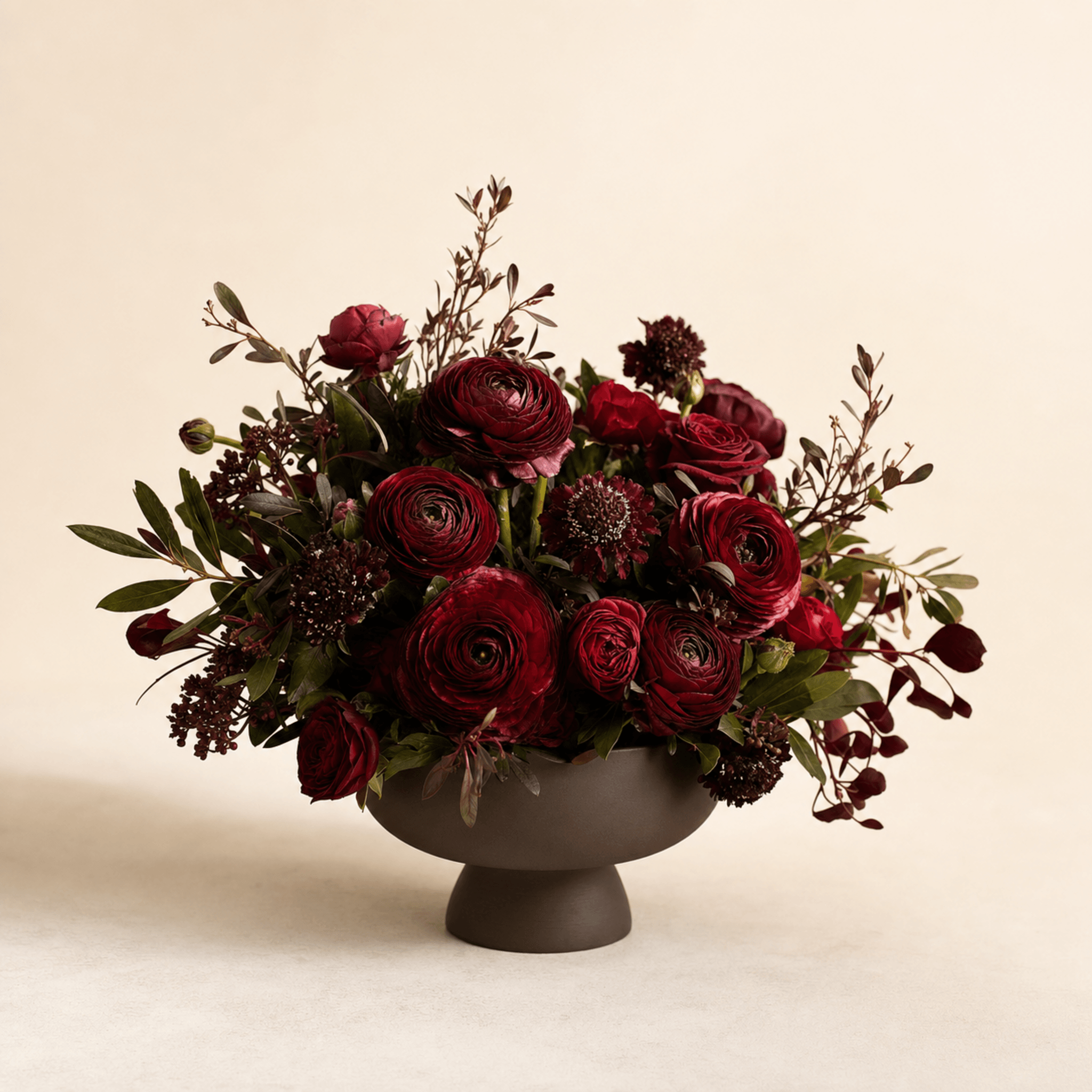 Deep red ranunculus, scabiosa, and roses with olive foliage in a dark footed ceramic bowl