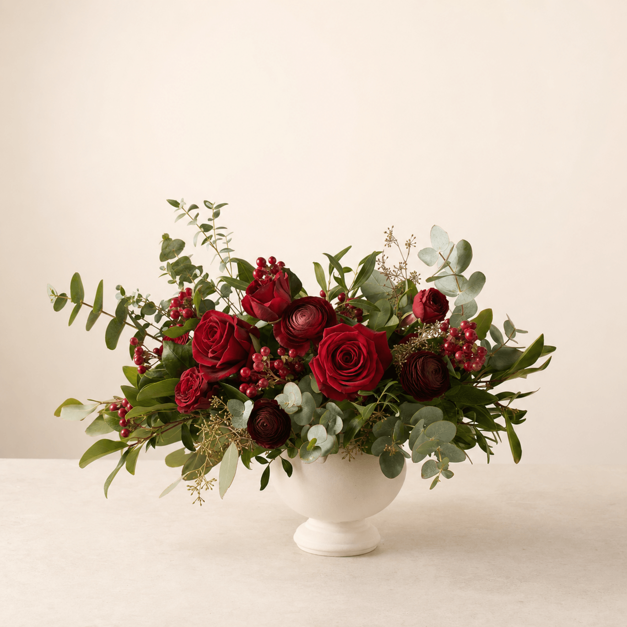 Red roses and ranunculus with eucalyptus and red berries in a white footed compote bowl