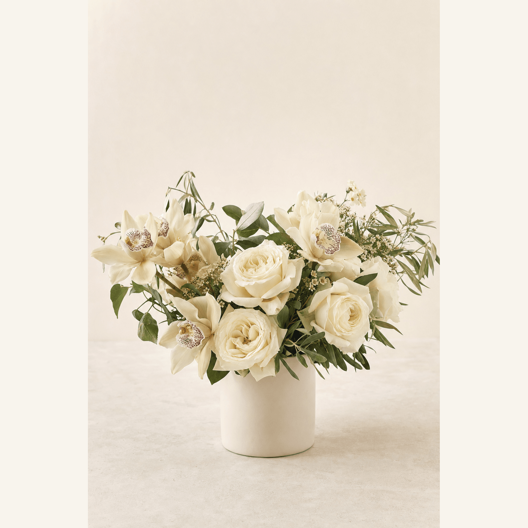 Cream roses and white cymbidium orchids with greenery in a white ceramic cylinder vase