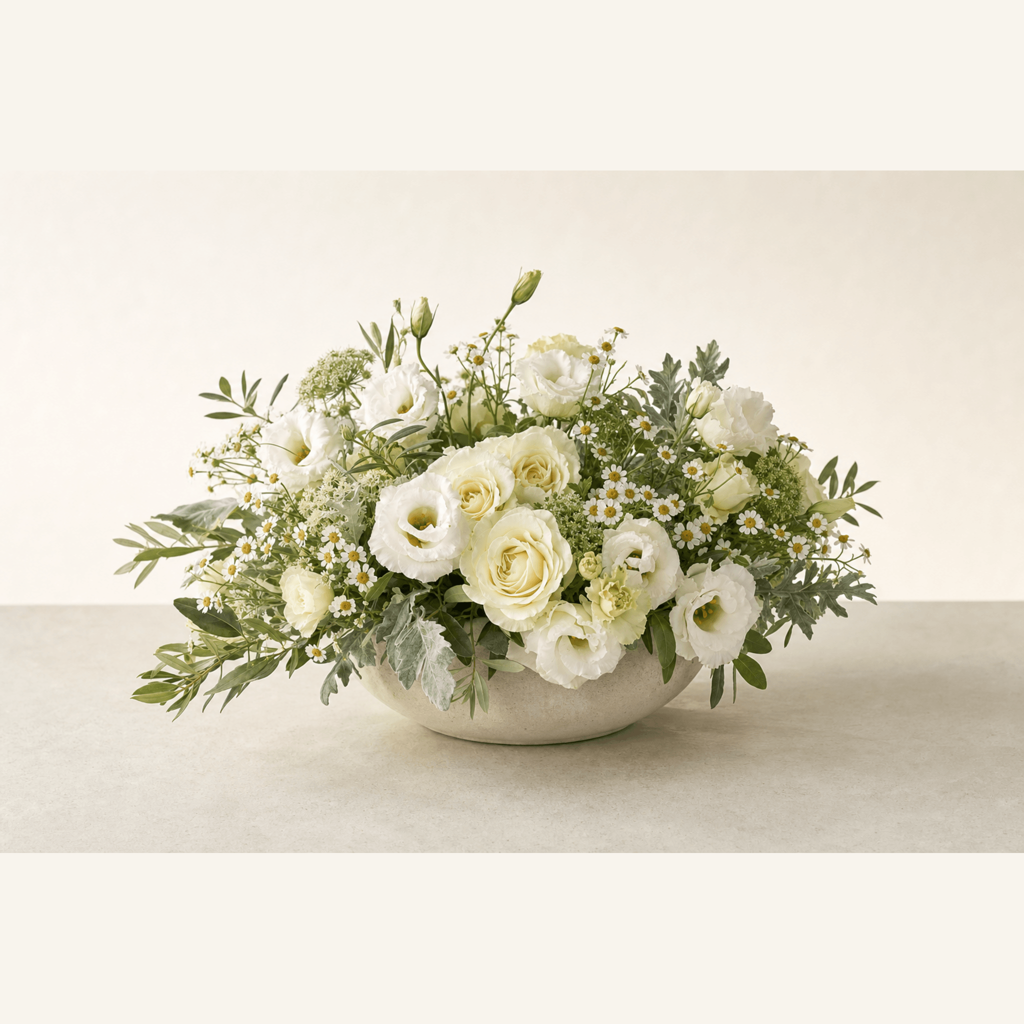 White roses, lisianthus, chamomile, and greenery arranged in a low stone bowl