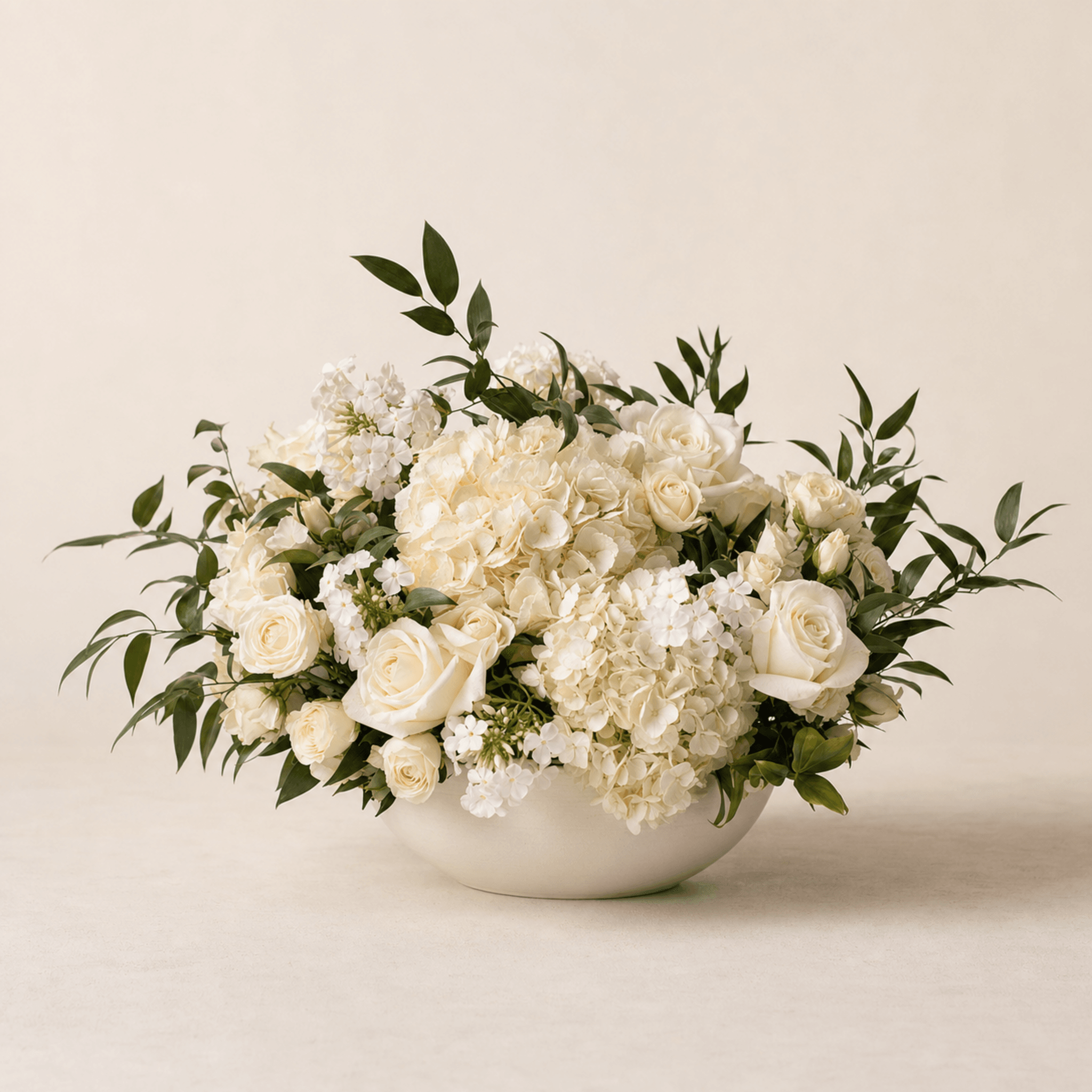 White roses, hydrangea, and stock with green foliage in a low cream ceramic bowl