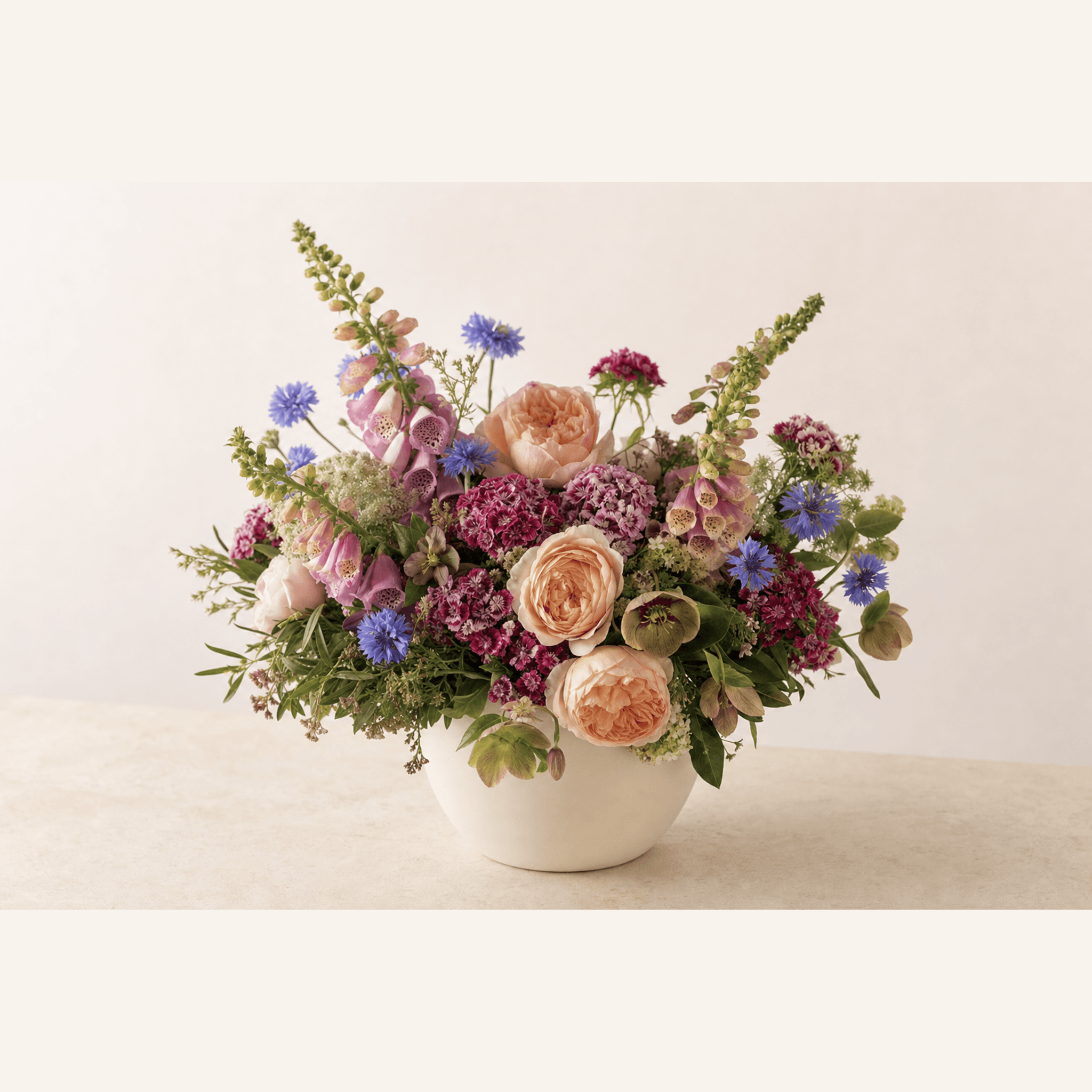 Peach garden roses, pink foxgloves, blue cornflowers, and magenta sweet William in a white ceramic bowl