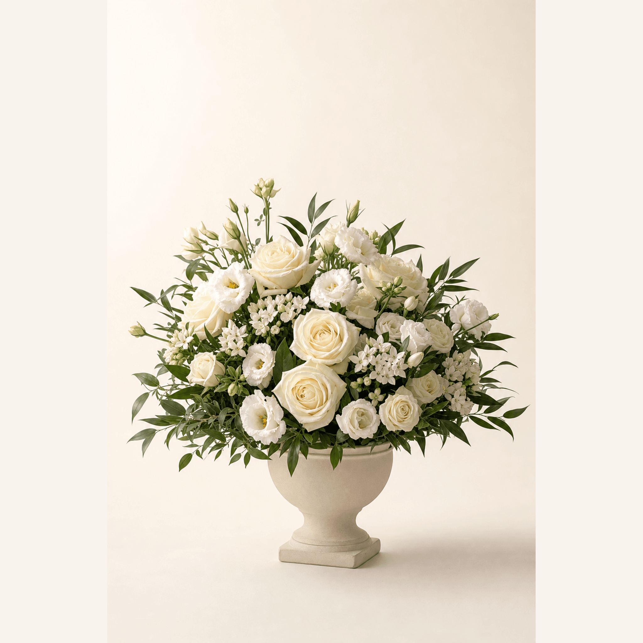 Cream roses, white lisianthus, and green foliage arranged in a cream footed urn vase