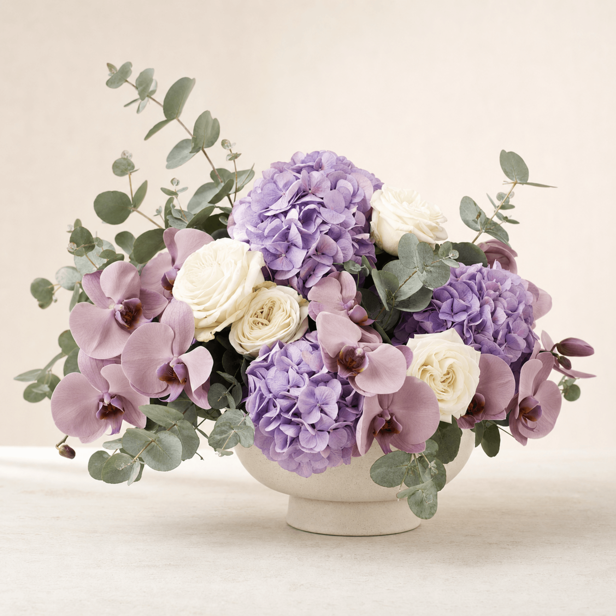 Purple hydrangeas, mauve orchids, white roses, and eucalyptus in a cream ceramic footed bowl