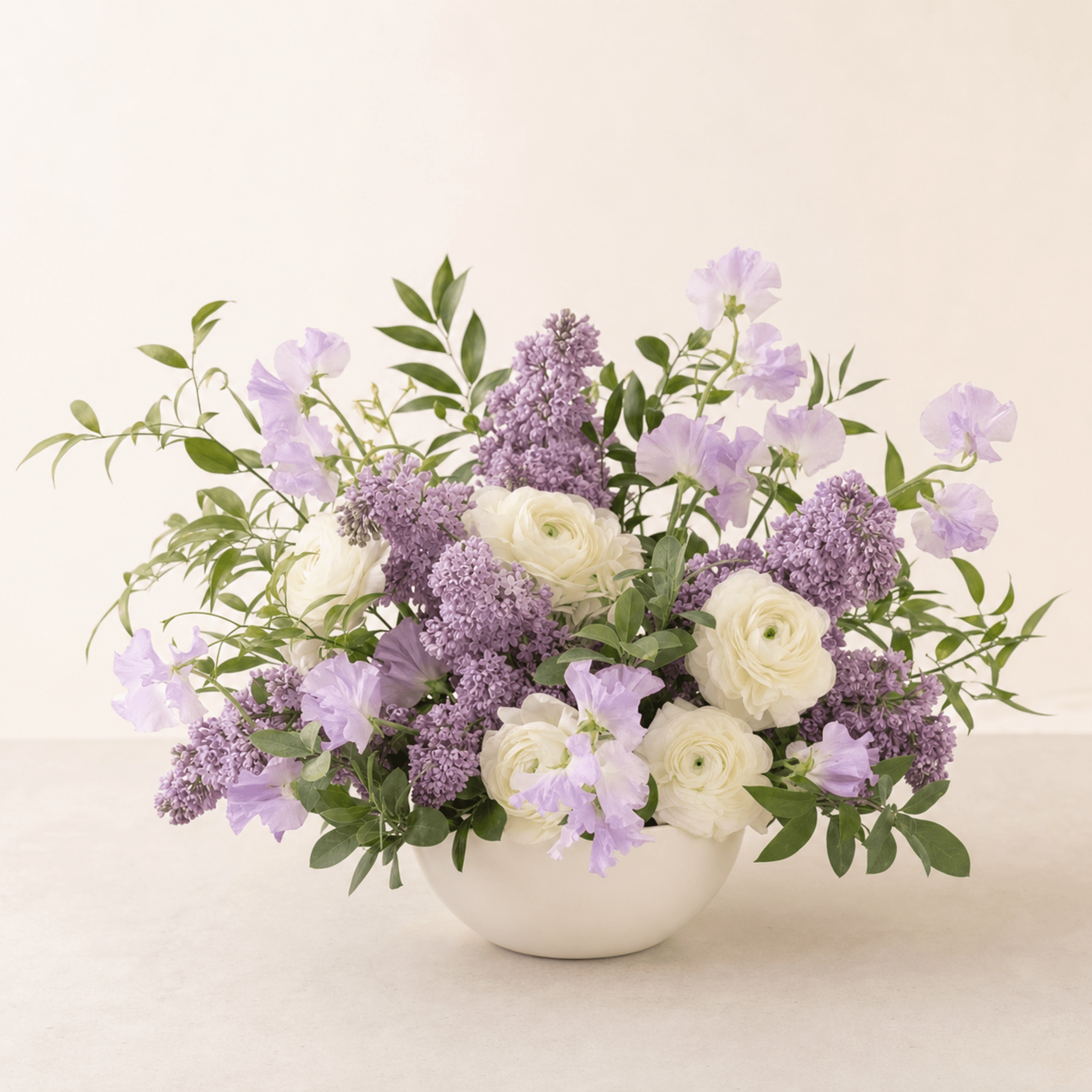 White ranunculus, purple lilacs, and lavender sweet peas in a white ceramic bowl