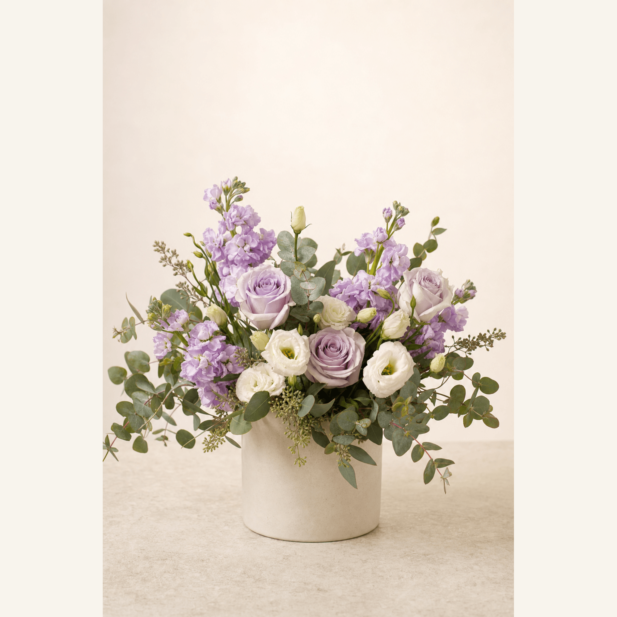 Lavender roses, white lisianthus, purple stock, and eucalyptus in a white ceramic pot