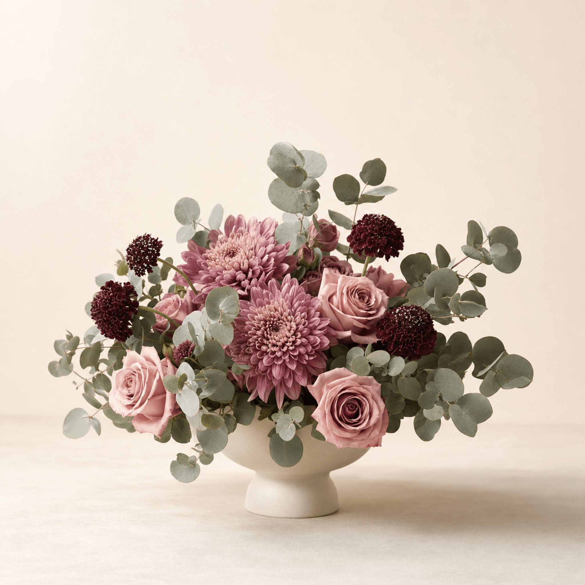 Mauve chrysanthemums, blush roses, burgundy scabiosa, and eucalyptus in a white footed ceramic bowl