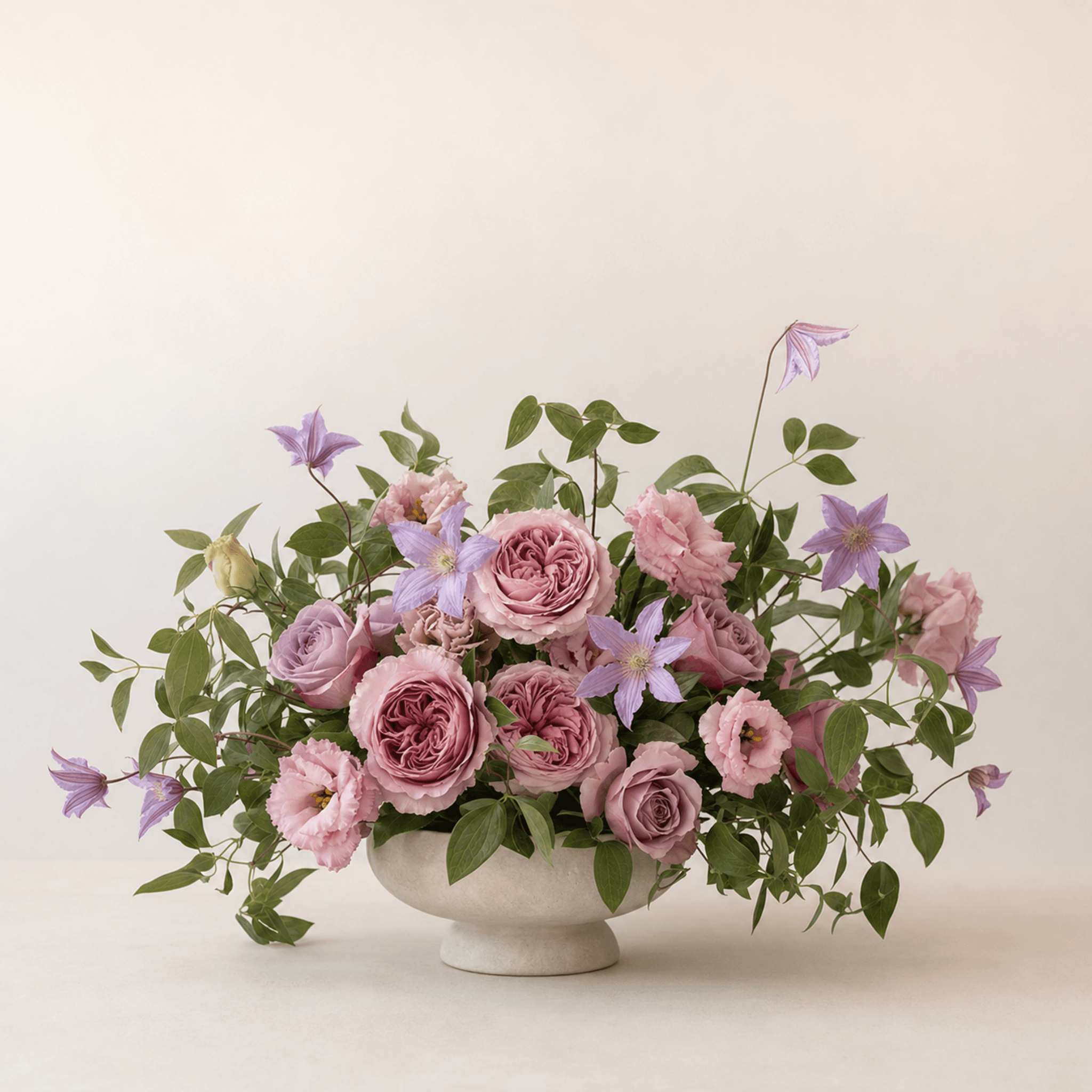 Pink garden roses, lisianthus, purple clematis, and trailing greenery in a stone footed bowl
