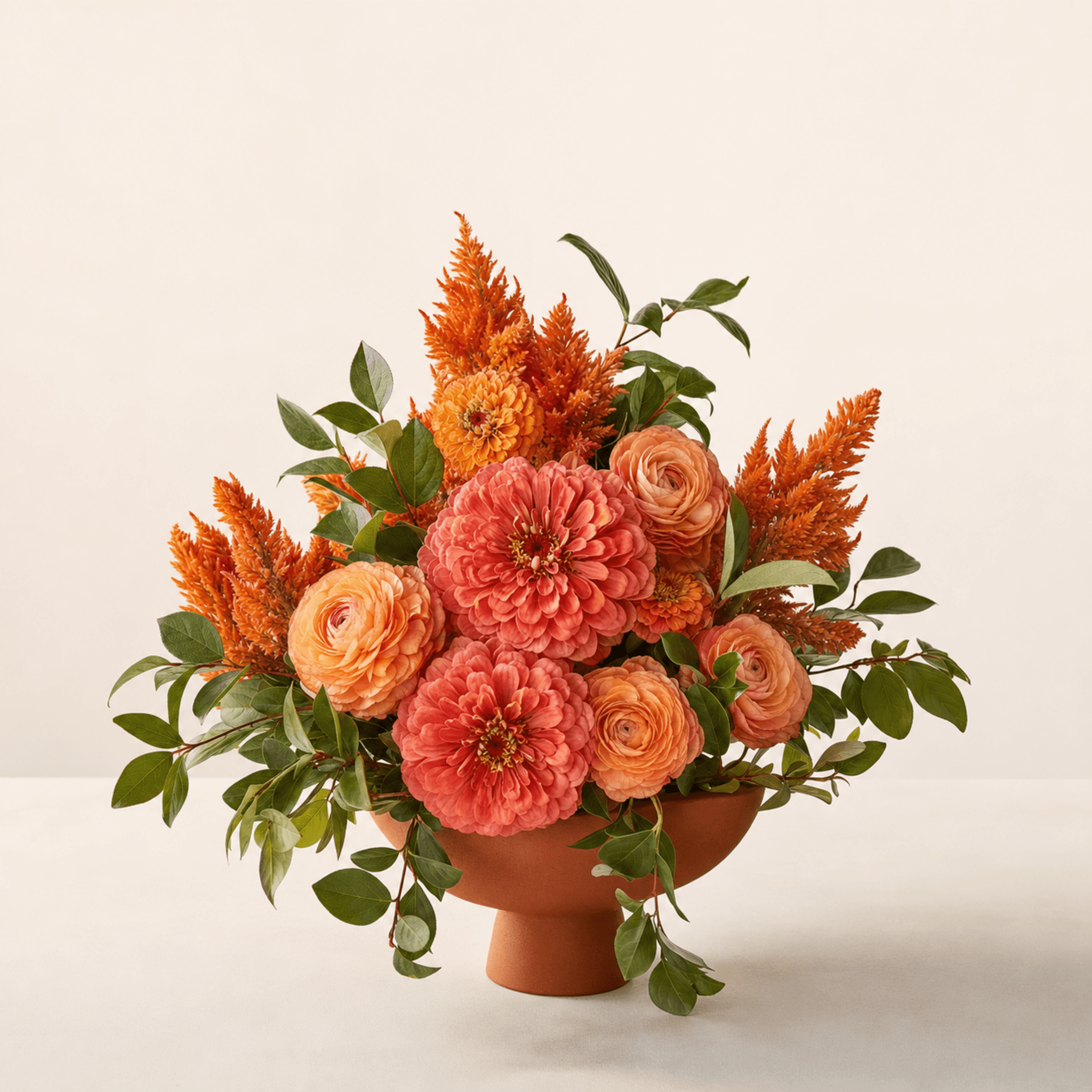 Coral zinnias, peach ranunculus, orange celosia, and trailing greenery in a terracotta footed bowl