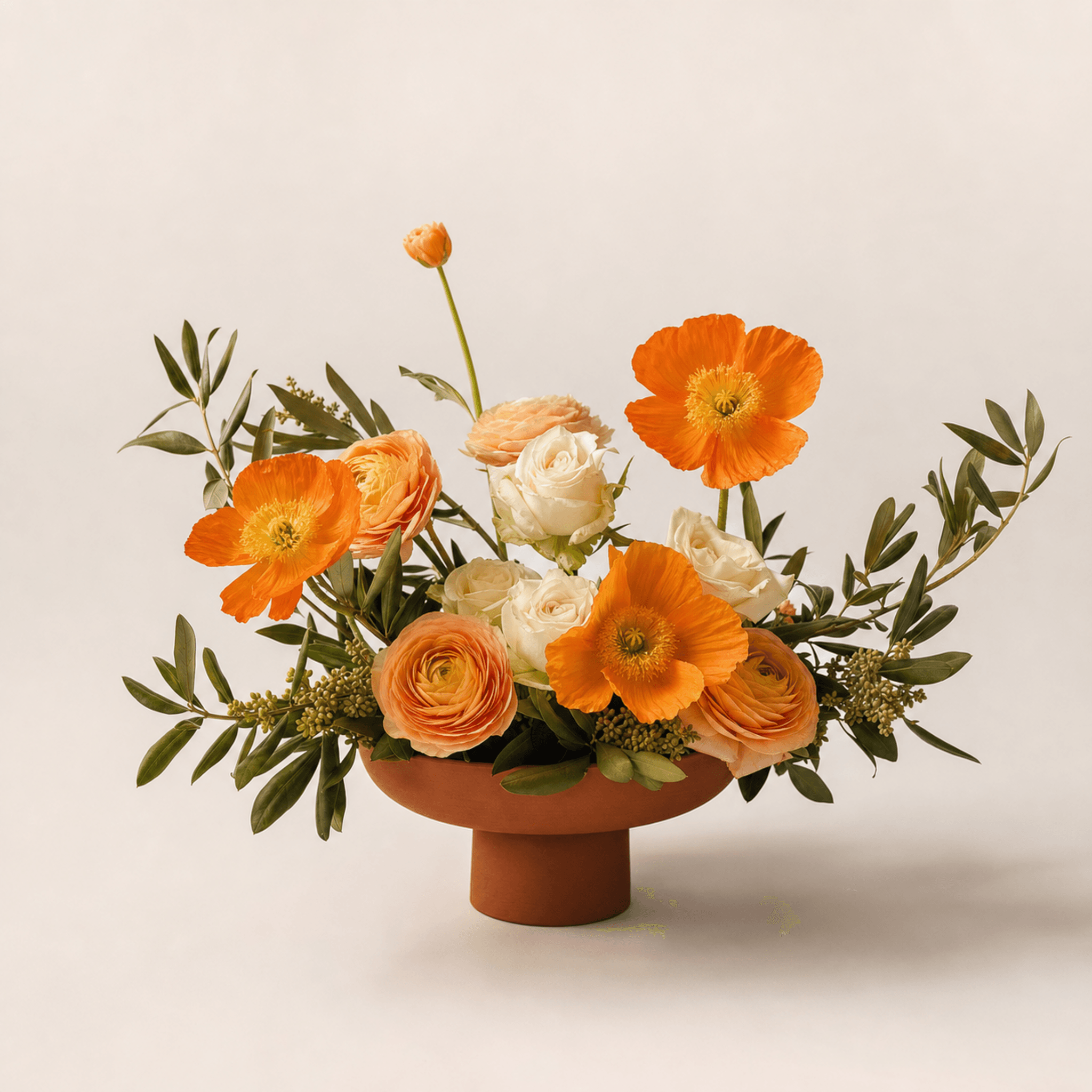 Orange poppies, peach ranunculus, white roses, and olive foliage in a terracotta footed bowl