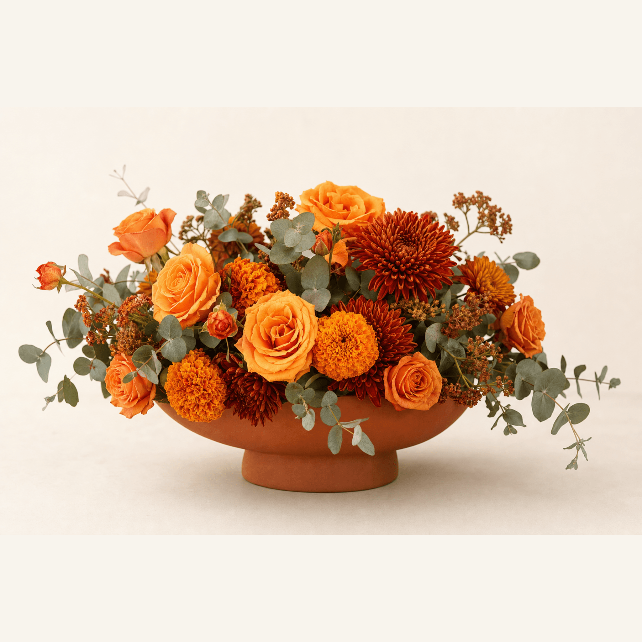 Orange roses, marigolds, rust chrysanthemums, and eucalyptus in a terracotta footed bowl