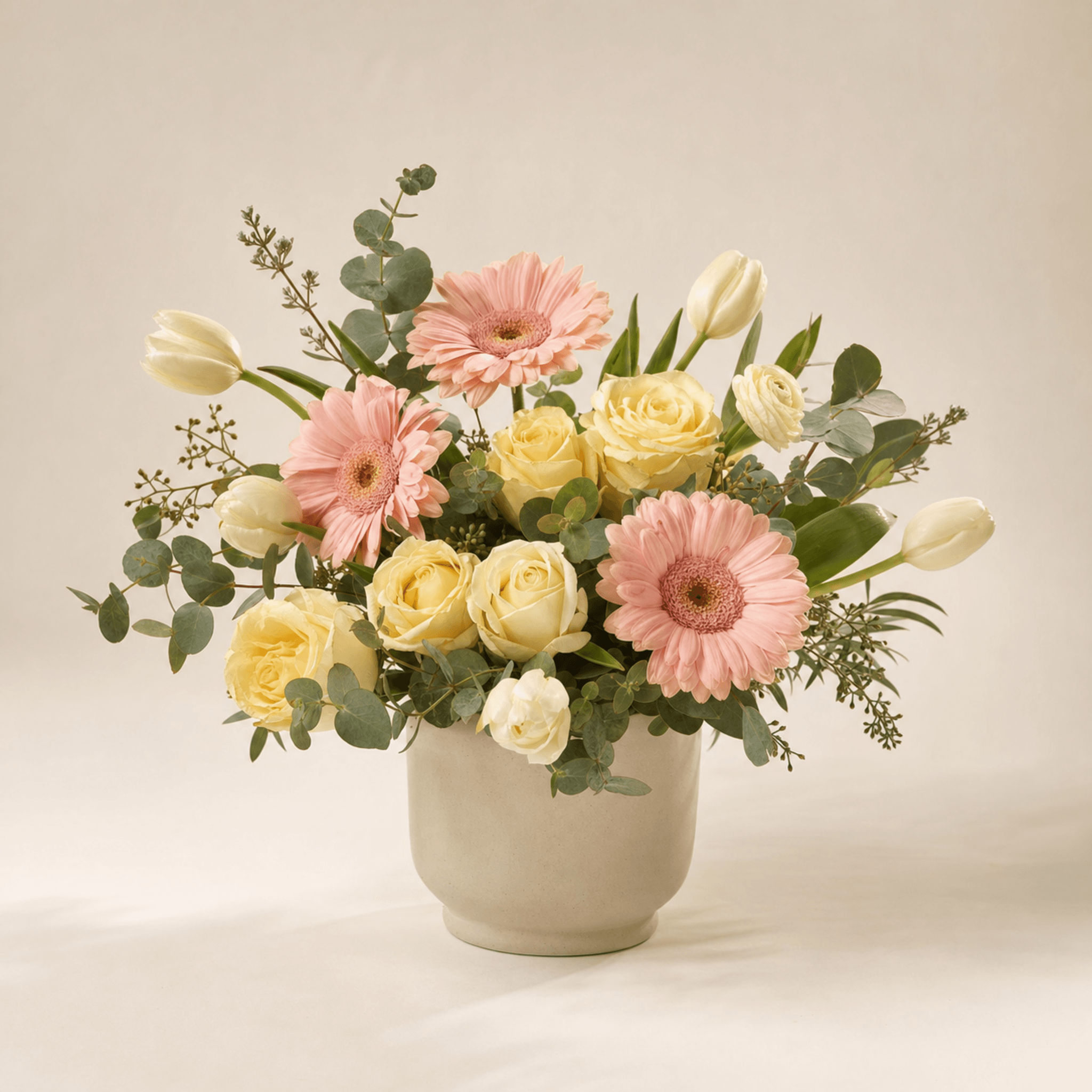 Pale yellow roses, pink gerberas, white tulips and eucalyptus in a cream ceramic pot