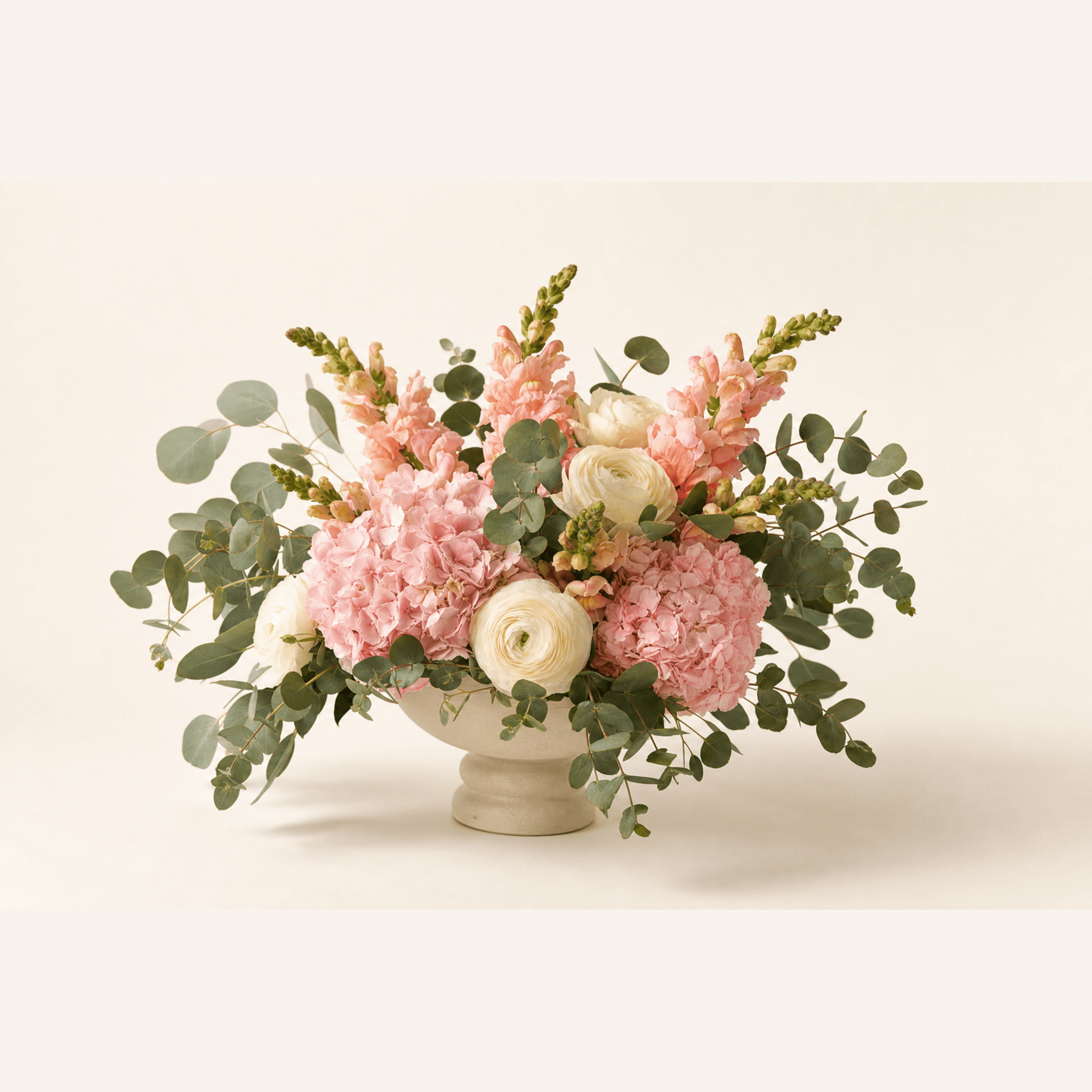 Pink hydrangeas, white ranunculus, and peach snapdragons with eucalyptus in a footed ceramic bowl