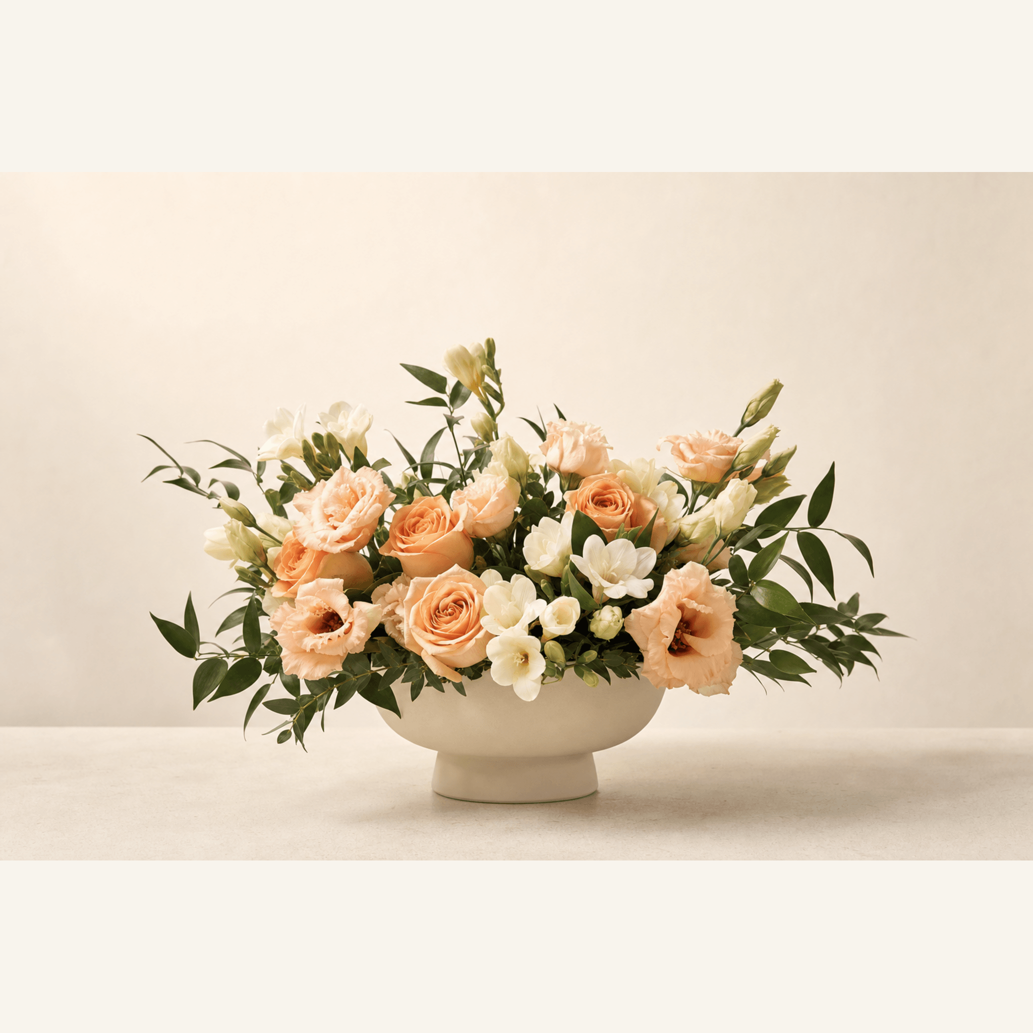 Peach roses, white freesia, and lisianthus with eucalyptus in a white footed ceramic bowl