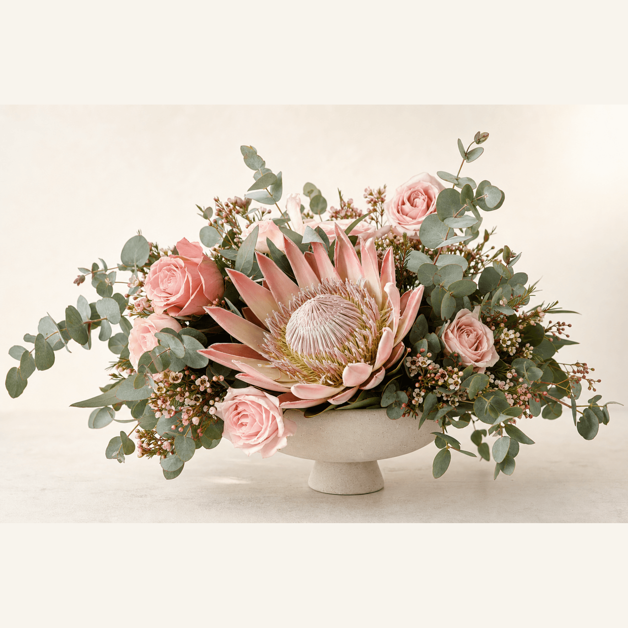 Blush king protea, pink roses, wax flower, and eucalyptus in a white footed bowl
