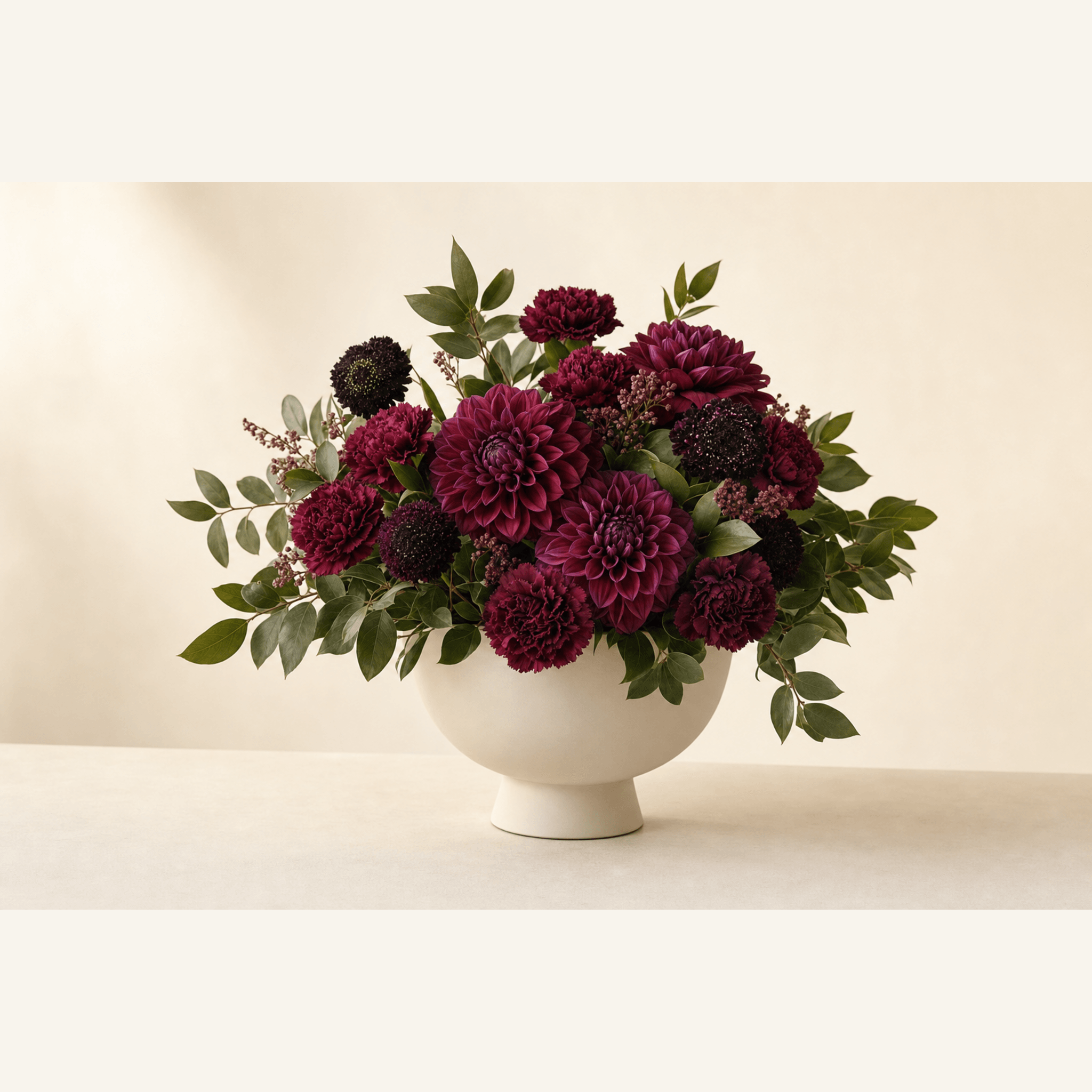 Deep burgundy dahlias, carnations, and scabiosa in a white footed ceramic bowl with eucalyptus