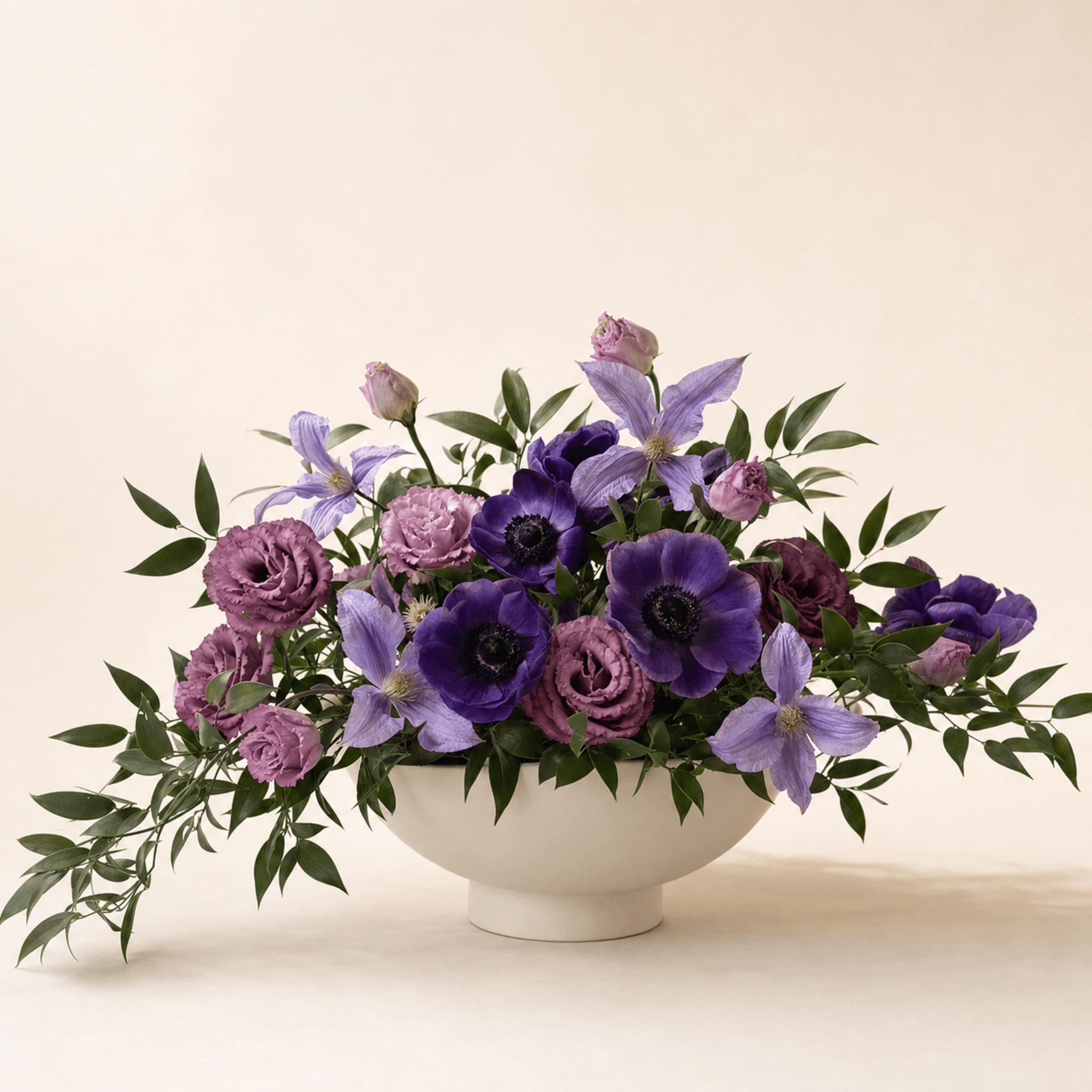Purple anemones, lavender clematis, and mauve lisianthus in a white footed ceramic bowl