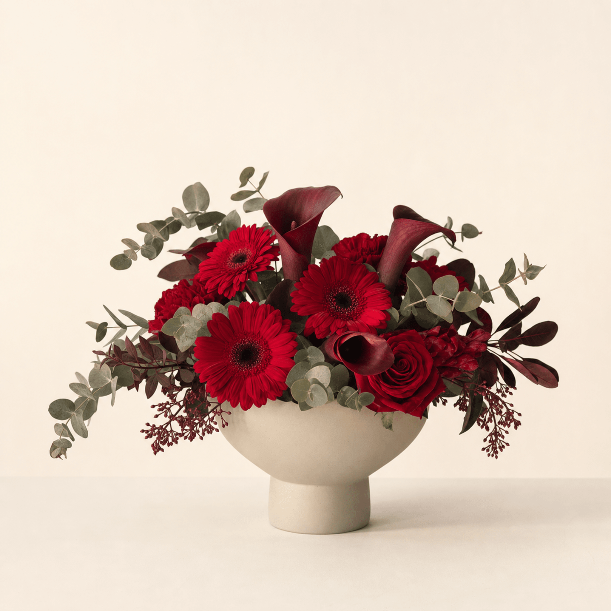 Red gerberas, dark calla lilies, red roses and eucalyptus in a white footed ceramic bowl