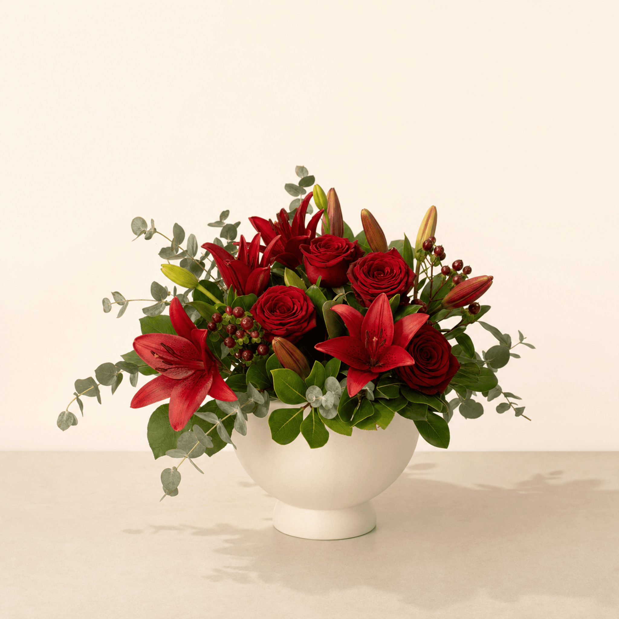 Red roses, asiatic lilies, hypericum berries and eucalyptus in a white footed ceramic bowl