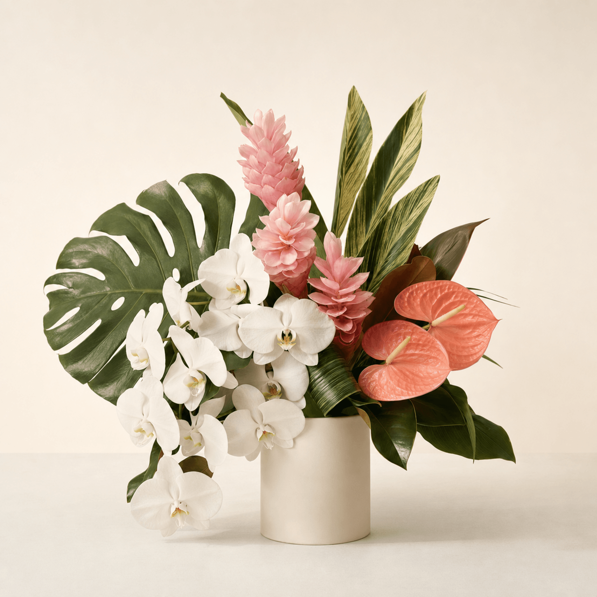 White phalaenopsis orchids, pink ginger, coral anthuriums, and monstera leaves in a cream ceramic cylinder vase