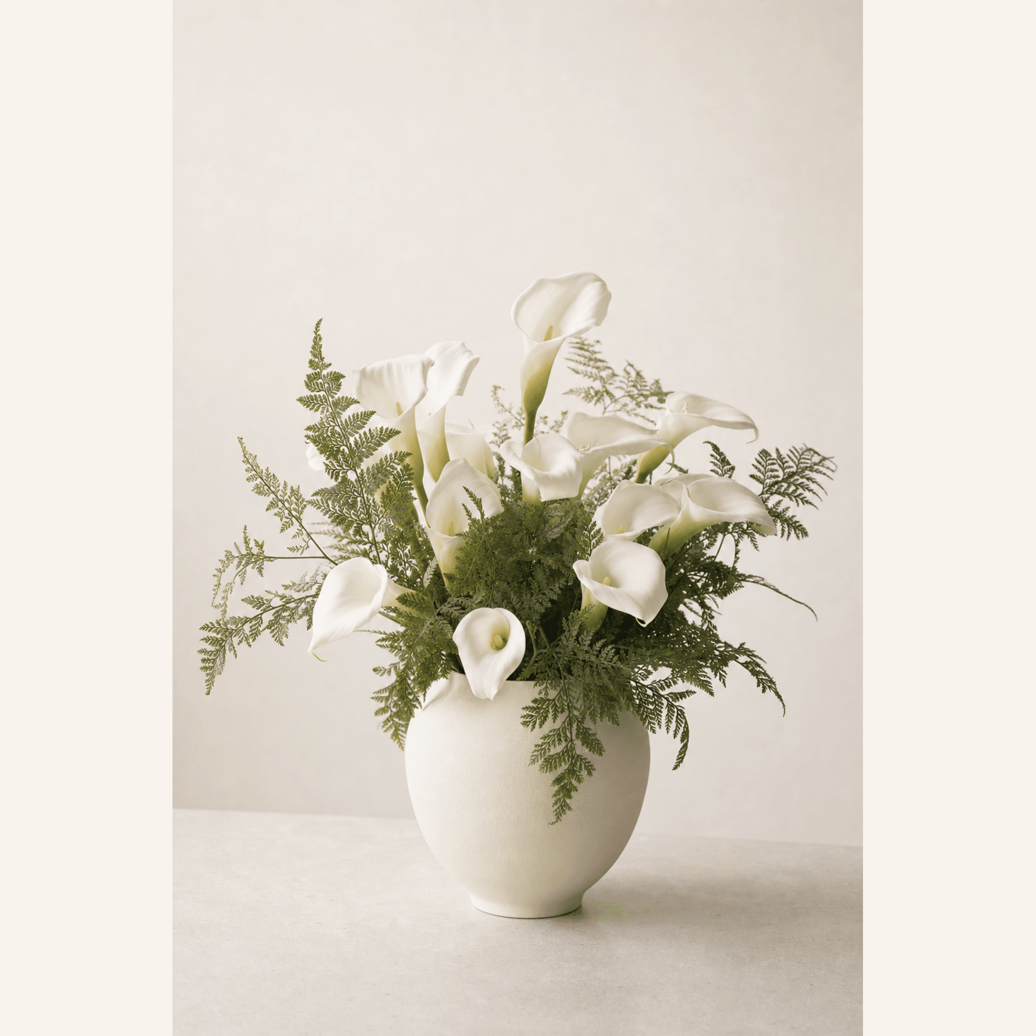 White calla lilies and green fern fronds arranged in a round cream ceramic vase