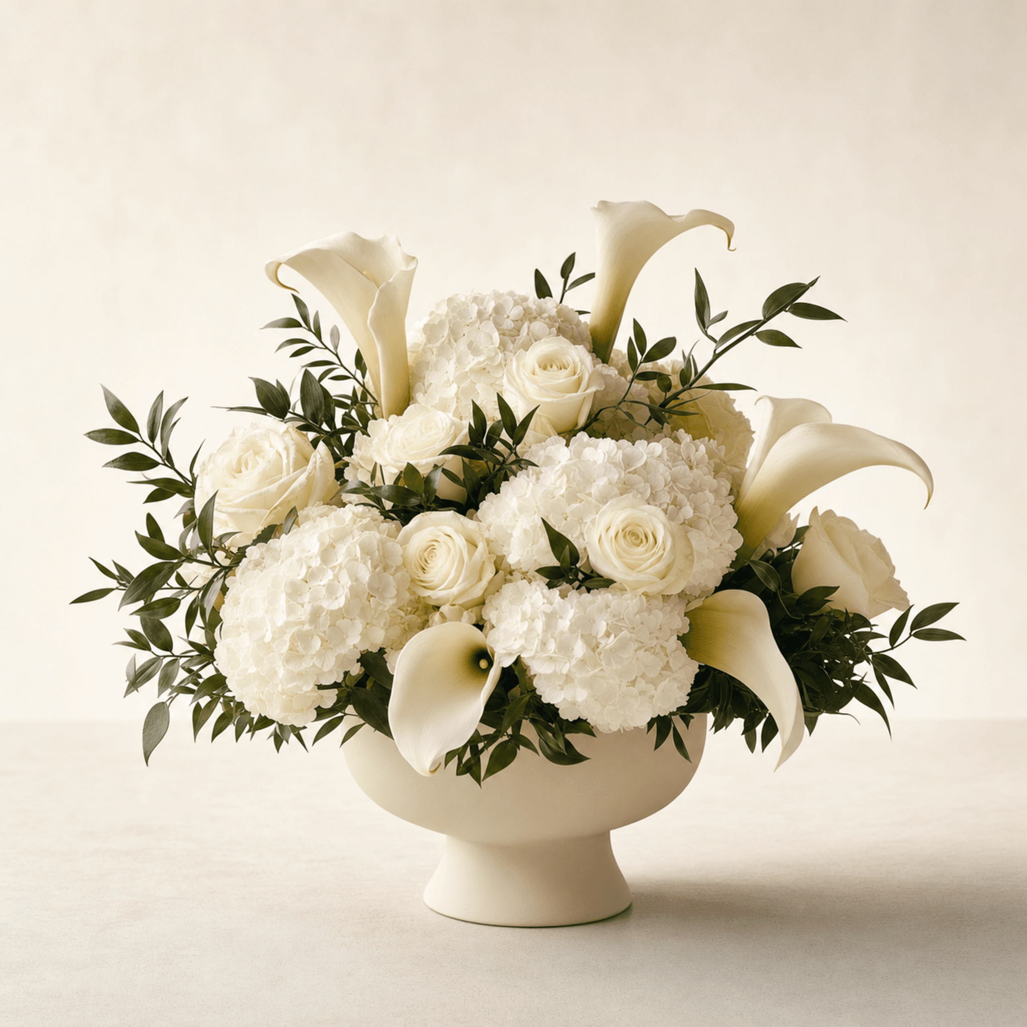 White calla lilies, roses, and hydrangea with greenery in a cream footed bowl