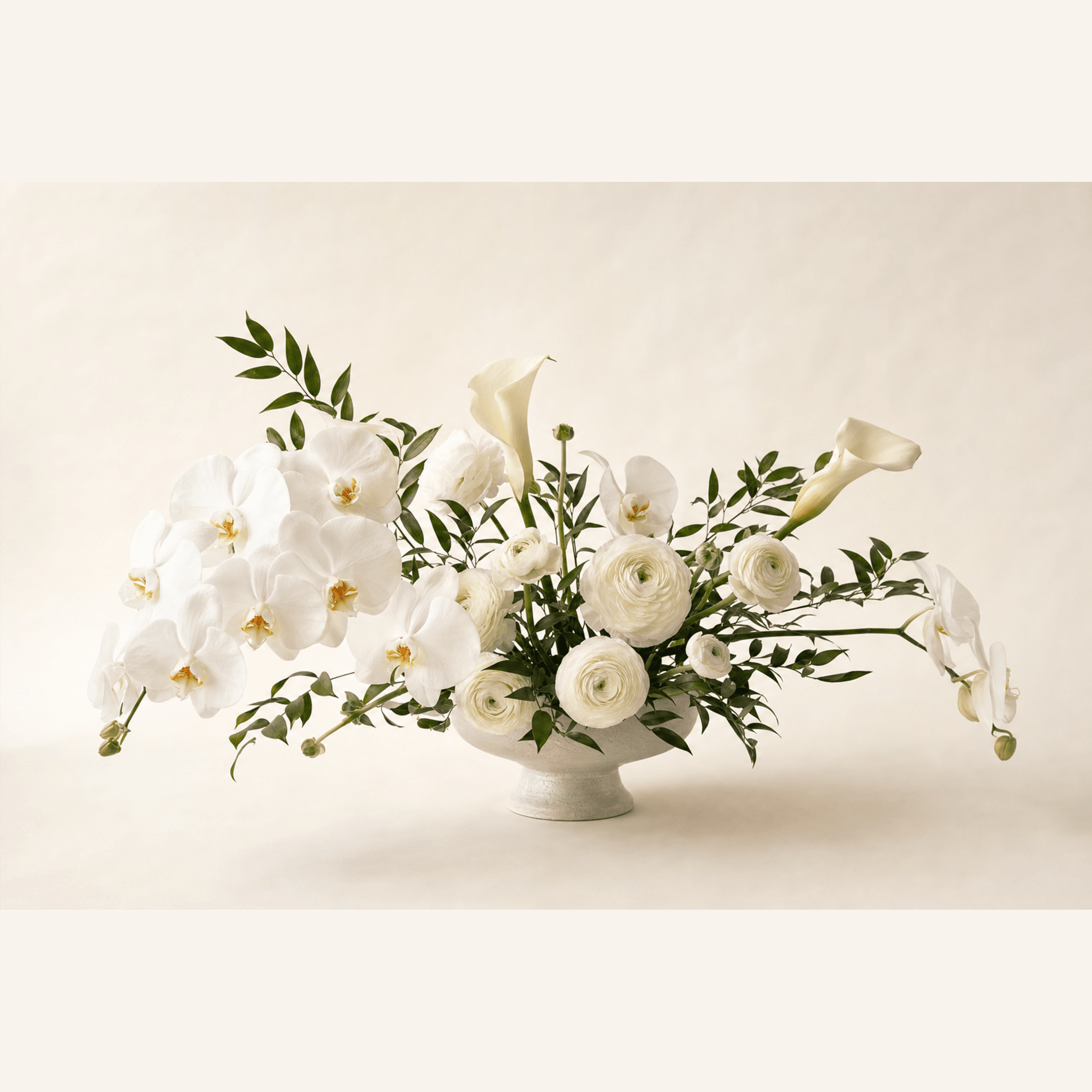 White orchids, ranunculus, and calla lilies with greenery in a white footed bowl