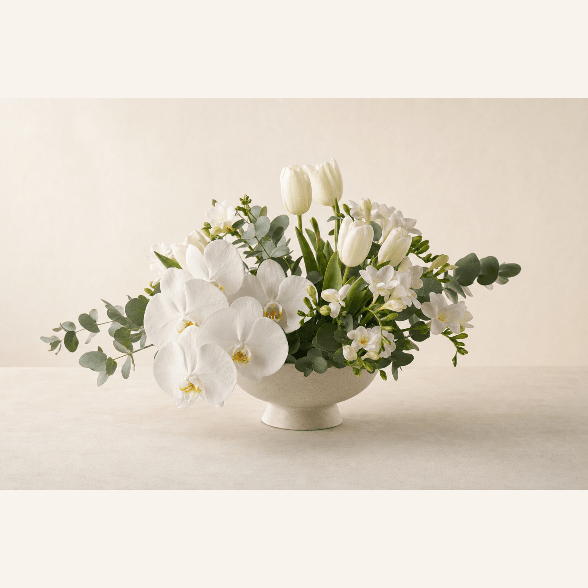 White phalaenopsis orchids, tulips, freesias, and eucalyptus in a cream footed ceramic bowl