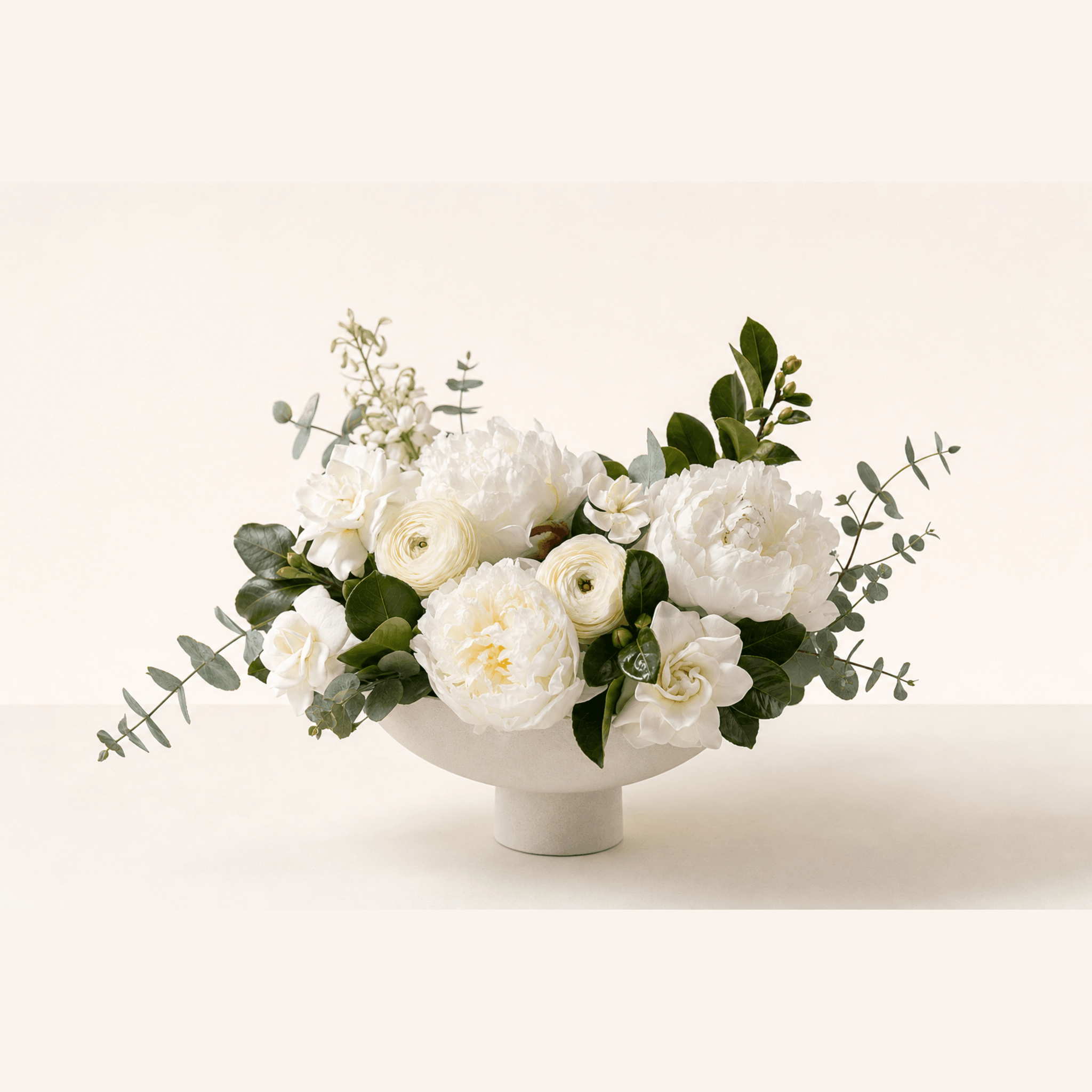 White peonies, ranunculus, gardenias, and eucalyptus in a white ceramic footed bowl