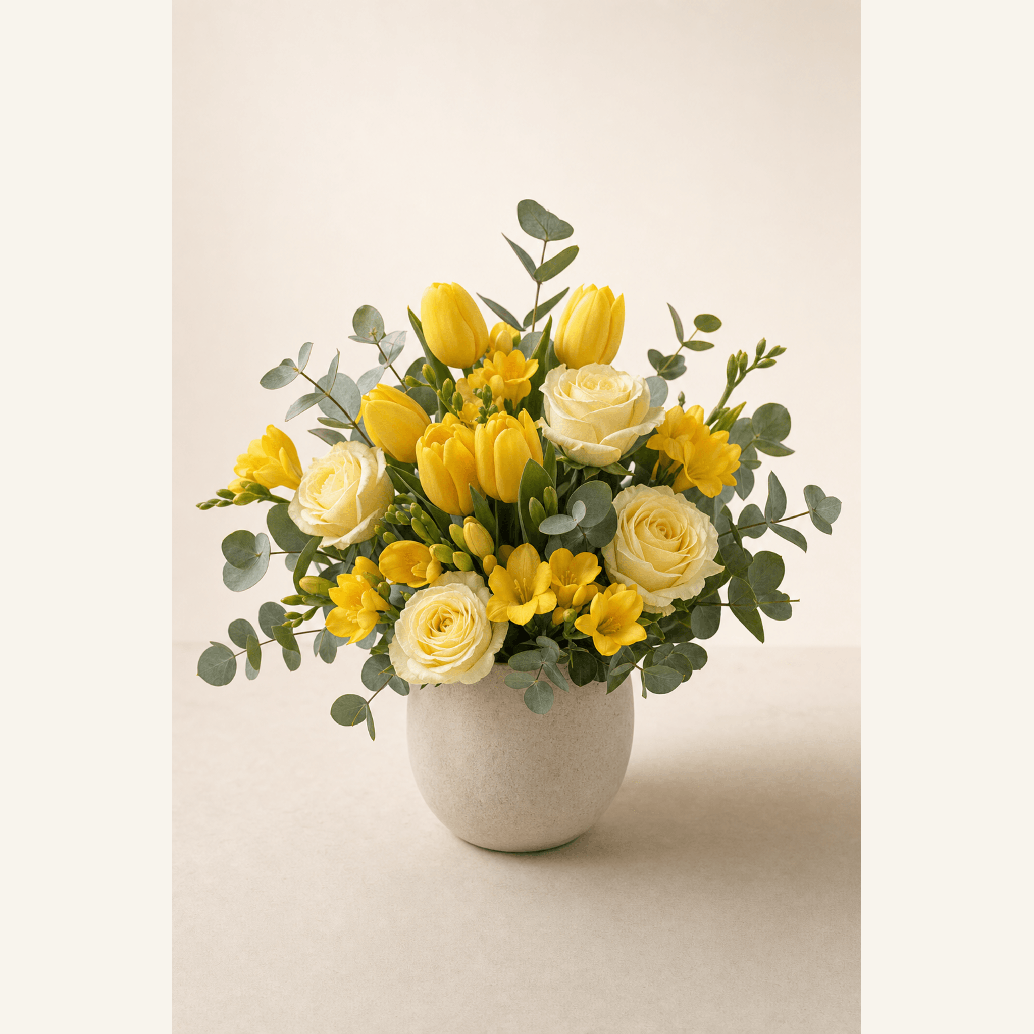 Yellow tulips, cream roses, yellow freesias and eucalyptus in a round stone ceramic pot