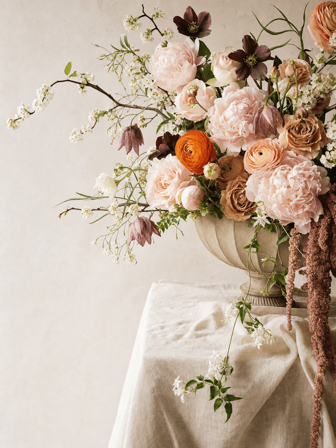 Blush peonies, orange and peach ranunculus, dark hellebores, cherry blossom branches and trailing amaranthus in a stone urn